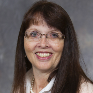 Professional headshot of Debbie Dean: A woman with long brown hair and glasses, smiling, wearing a white blouse and pearl necklace, in front of a gray background.