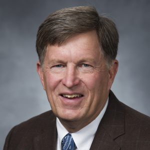 Professional headshot of Chris Crowe: A smiling older man with short brown hair wearing a dark suit, white shirt, and patterned blue tie, posed against a gray background.
