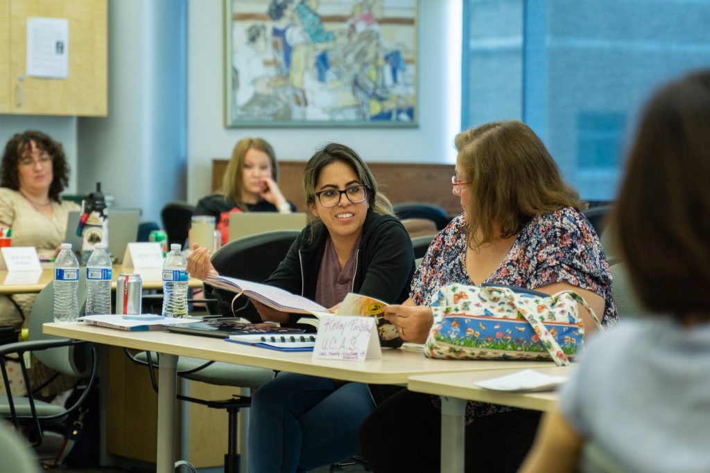 Two women sit at a classroom table having a conversation, with open notebooks and papers in front of them. Other people are seated in the background, and artwork hangs on the wall.