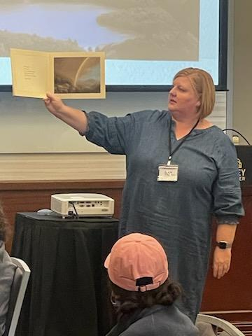 A woman stands in front of a classroom, holding up an open book with a rainbow photo, showing it to an audience. She wears a blue dress and name badge. People are seated facing her, one wearing a pink cap.