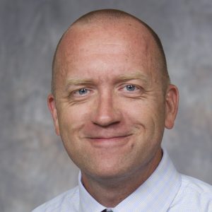 Professional headshot of Jon Ostensen: A man with blue eyes and a shaved head smiles at the camera. He is wearing a light-colored, collared shirt. The background is softly blurred in neutral tones.