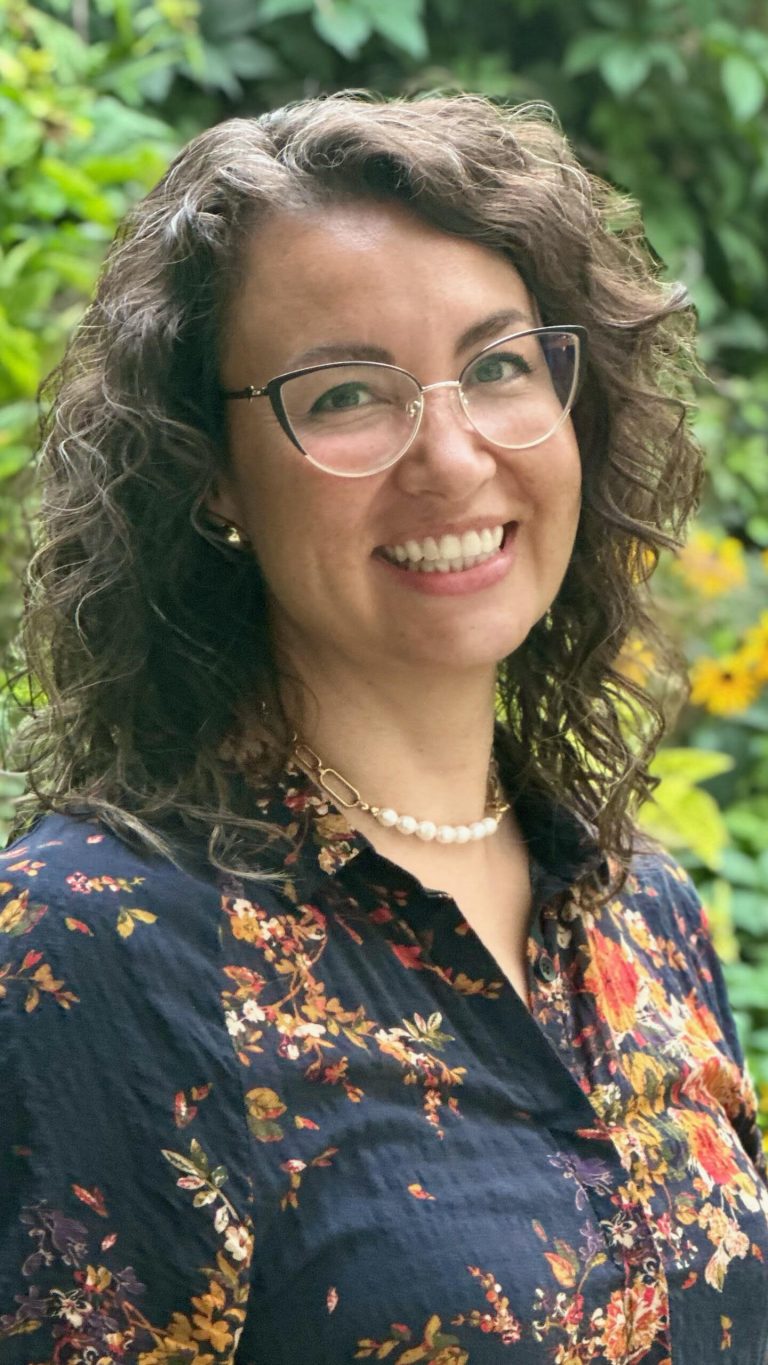 Professional headshot of Kristie Jolley: A woman with curly brown hair and glasses smiles outdoors. She is wearing a dark floral blouse, pearl necklace, and stud earrings, with greenery and yellow flowers in the blurred background.