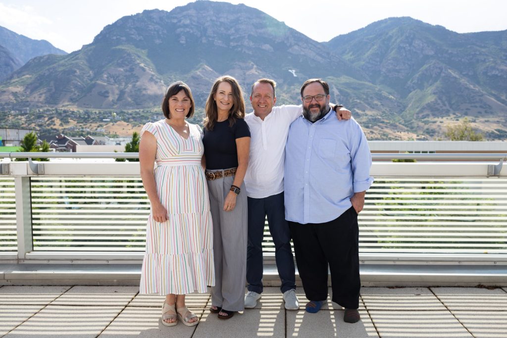 Four adults stand close together, smiling on a sunny balcony with mountains and greenery in the background. Two women are on the left in light clothing, two men on the right in shirts and pants.