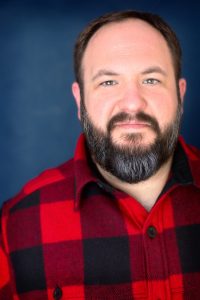 Professional headshot of Joseph Wiederhold: A bearded man wearing a red and black plaid shirt stands against a dark blue background, looking directly at the camera with a neutral expression.