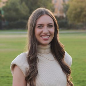 Professional photograph of Leah Parks: A woman with long brown hair and a cream-colored sleeveless turtleneck top smiles while standing outdoors on a grassy field, with trees and buildings blurred in the background.