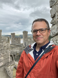 Professional photograph of Brian Jackson: A man wearing glasses and an orange jacket smiles while taking a selfie at a historic stone castle with tall towers and cloudy skies in the background.