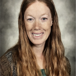 A woman with long, wavy red hair smiles at the camera. She is wearing a green top and a brown patterned sweater, and is posed against a gray, softly blurred background.