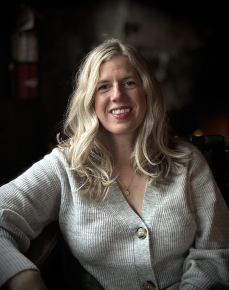 A woman with long blonde hair, wearing a light gray cardigan with large buttons and a gold necklace, smiles while sitting indoors against a dark, softly lit background.