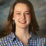A young woman with light skin and straight brown hair smiles at the camera. She is wearing silver earrings, a necklace, and a blue and white checkered shirt, with a cloudy background behind her.