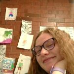 A person with long curly hair and glasses smiles at the camera, resting their chin on their hand. Behind them are colorful drawings, papers, and charts pinned to a brick wall.
