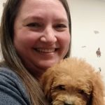 A smiling woman with long brown hair holds a fluffy, light brown puppy close to her chest, both looking toward the camera. The background is indoors with faint wall decorations visible.