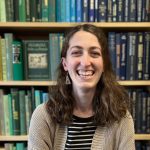 A person with curly brown hair, wearing a striped shirt and beige cardigan, is smiling broadly while sitting in front of bookshelves filled with colorful books.