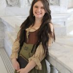A young woman with long brown hair, wearing a medieval-style dress in beige and green, stands smiling and holding a book in a marble hall with ornate wall details.