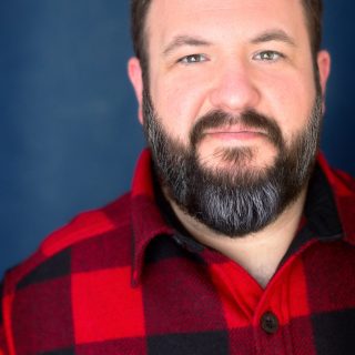Professional headshot of Joseph Wiederhold: A bearded man wearing a red and black plaid shirt stands against a dark blue background, looking directly at the camera with a neutral expression.
